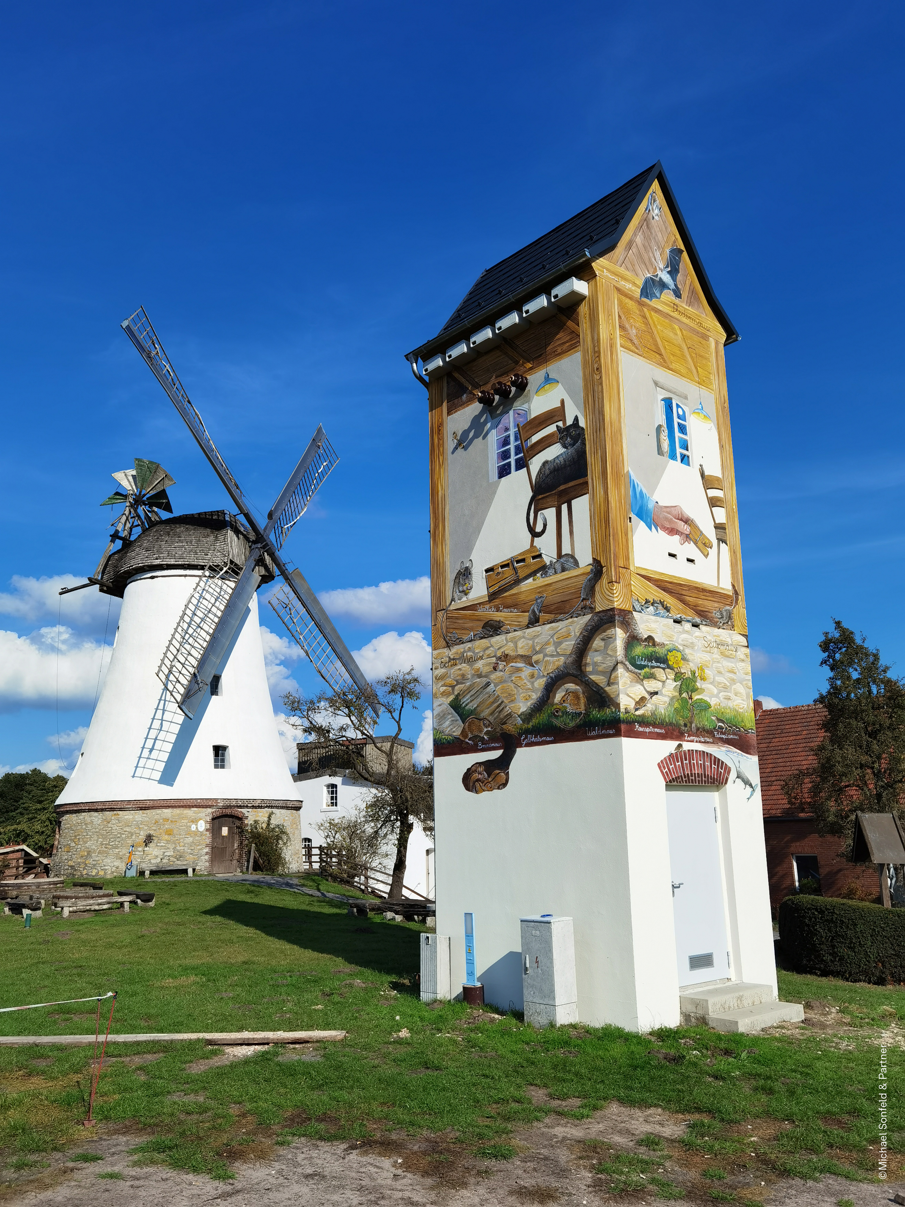 Im Vordergrund steht ein Trafoturm, der bemalt ist. Im Hintergrund ist eine Mühle zu sehen. 