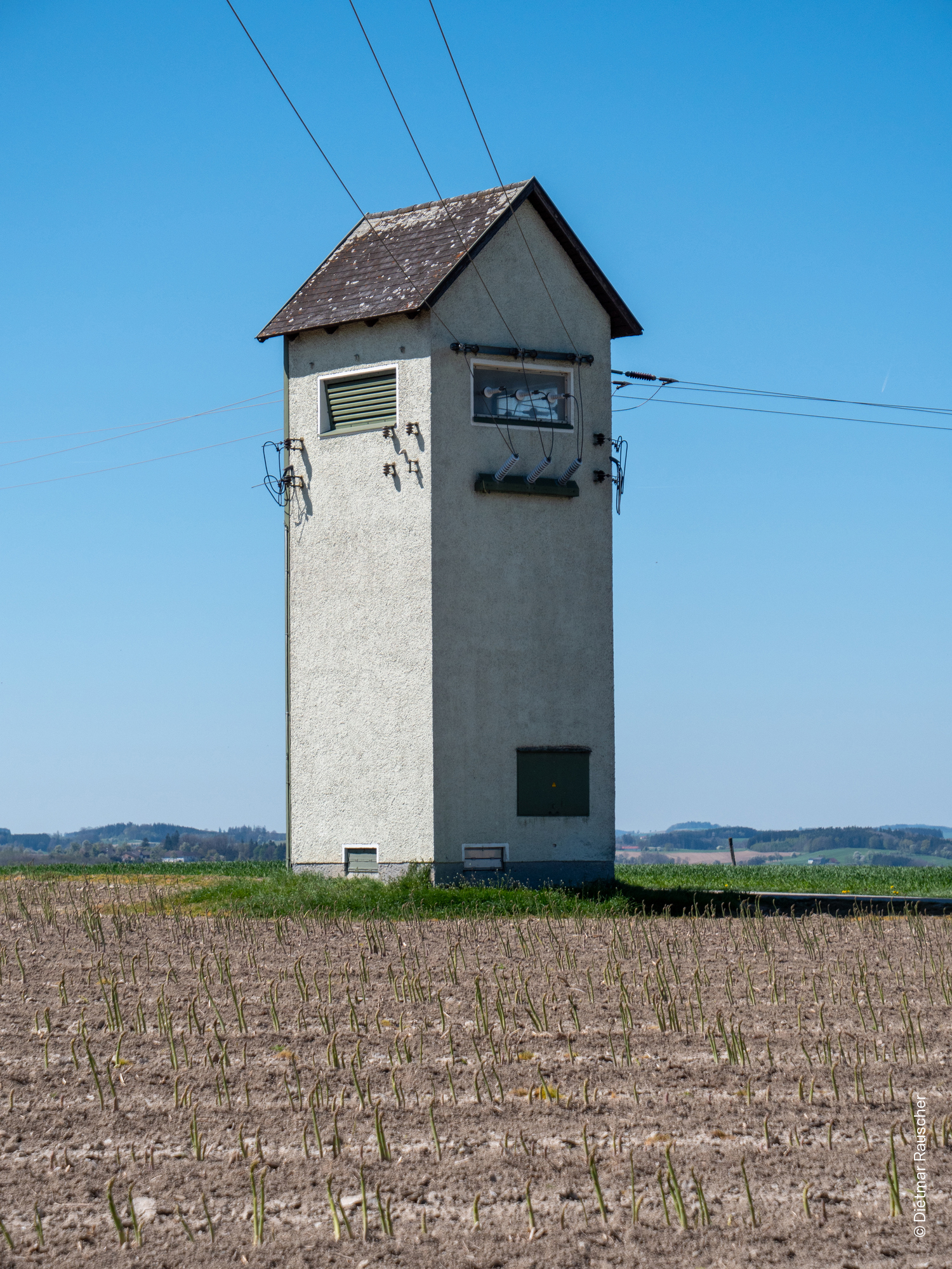 Alter Trafoturm auf freiem Feld unter blauem Himmel mit umliegender junger Vegetation.