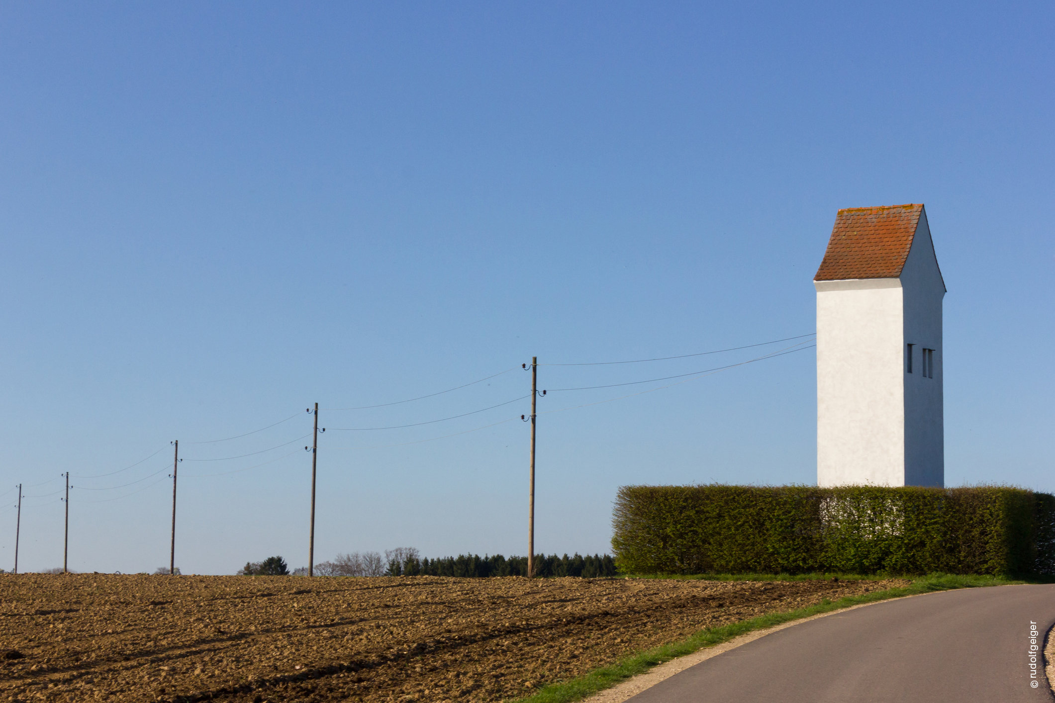 Weiß verputzter Trafoturm mit rotem Ziegeldach an einem Feldweg neben einer Hecke und Strommasten.