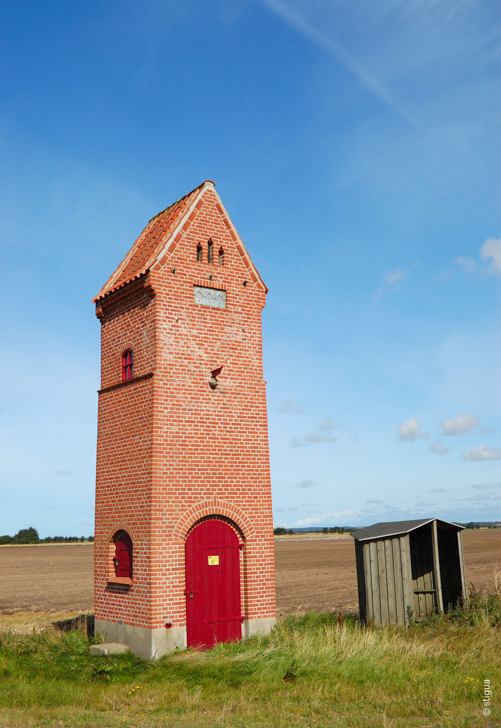Roter Backstein-Trafoturm mit roter Tür und Fenstern auf freiem Feld unter blauem Himmel.